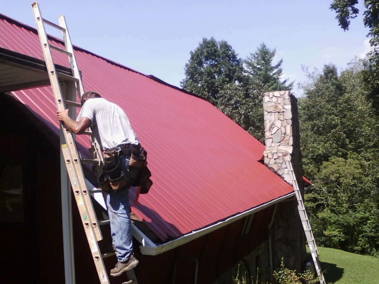 Roofer on a ladder beside a red metal roof and stone chimney — Soffit & Fascia Repair by Roofing Brothers in Toston