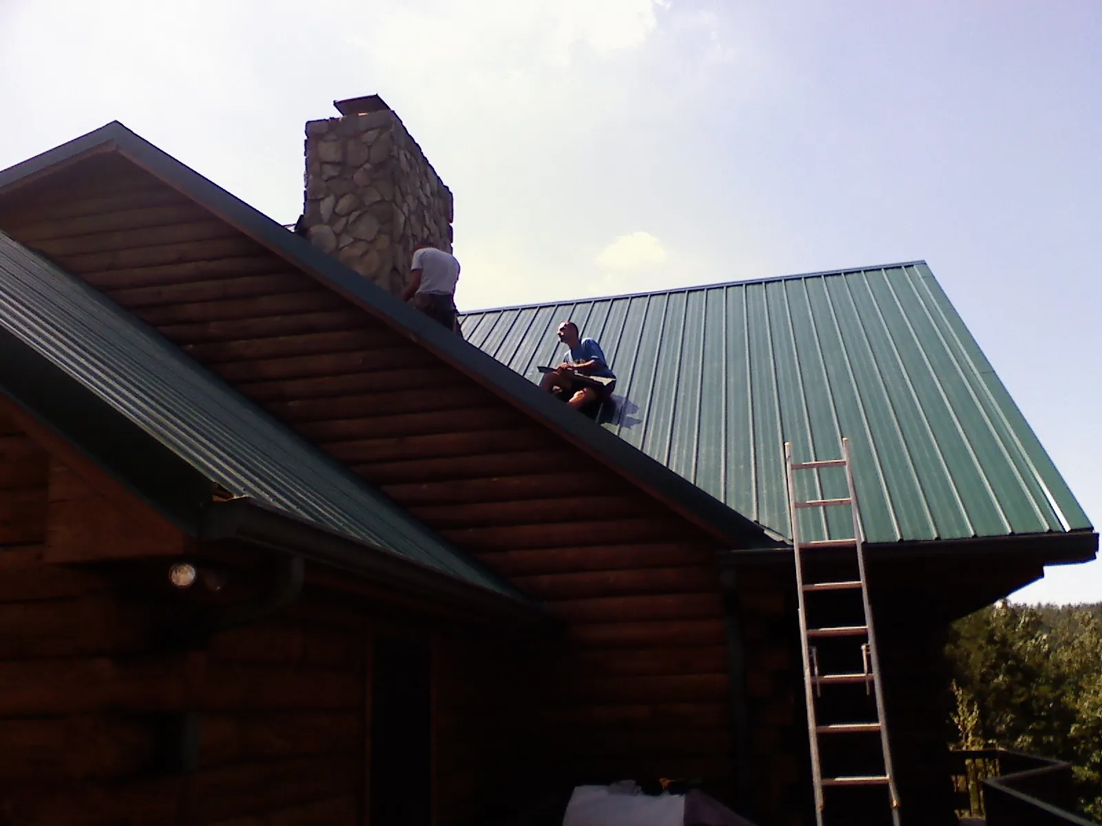 Roofers installing green metal panels beside a stone chimney on a log home in Toston
